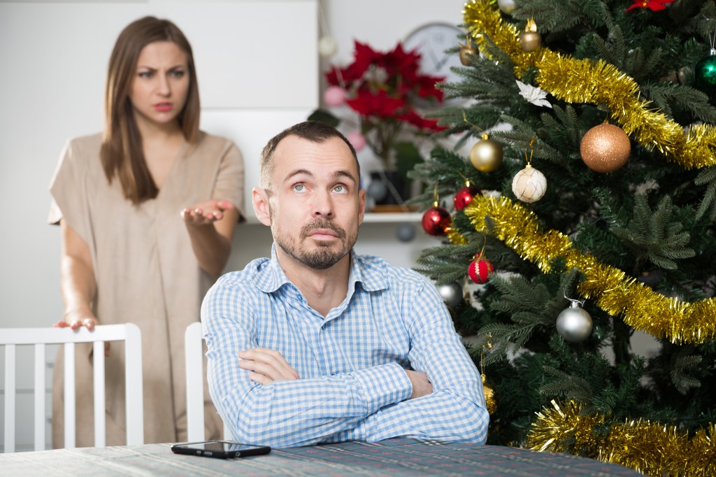 Frustrated man sitting at home table near Christmas tree with unhappy woman behind him during Christmas