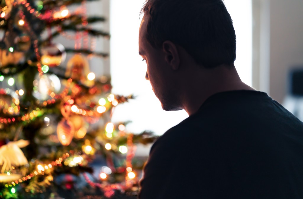 Depressed man addressing financial stress at Christmas standing alone in front of a festively decorated tree in a winter home.